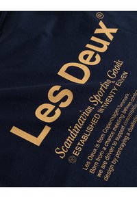 Námorní bavlněná tkanina s velkým zlatým textem, který zobrazuje název značky "Les Deux" a další text o skandinávských sportovních potřebách.