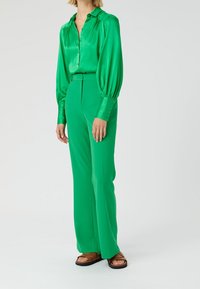 Blusa di raso verde con polsi abbottonati e maniche ampie, abbinata a pantaloni a vita alta coordinati. Indossata con sandali piatti marroni.