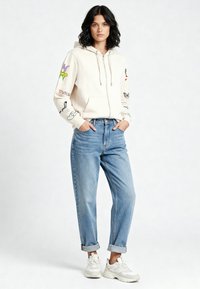 Kremasti hooded duks s šarenim vezom na rukavima, uparan s trapericama svijetloplave boje s podvrnutim rubom i bijelim tenisicama.