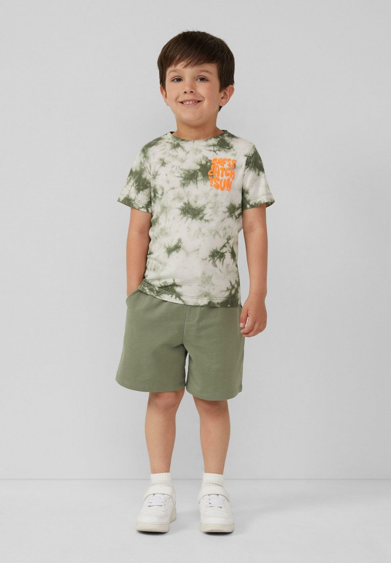 Groene tie-dye t-shirt met oranje tekst, gecombineerd met olijfgroene shorts. Kind draagt witte sneakers en sokken. Eenvoudig en casual ontwerp.