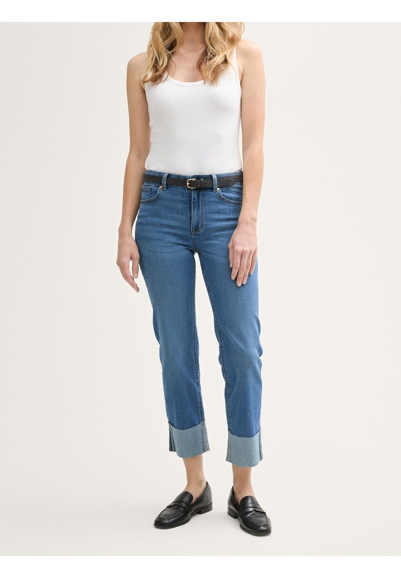 Femme portant un débardeur blanc, un jean bleu retroussé avec une ceinture noire et des mocassins noirs, debout devant un fond uni.
