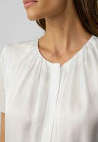 Weiße Bluse mit Rundhalsausschnitt, plissierten Details und kurzen Ärmeln. Hergestellt aus einem glatten, leichten Stoff mit einem subtilen Glanz.