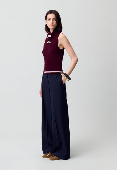 Top sin mangas de punto burdeos con cuello alto y rayas blancas; combinado con pantalones de pinzas de pierna ancha en azul marino y zapatos de gamuza marrón.
