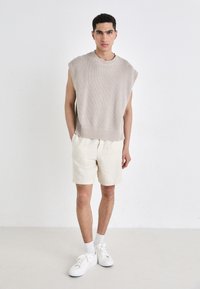 Beige strikket ermeløs genser med ribbestrikket kant, sammen med lysebeige linshorts og hvite joggesko, med hvite sokker.