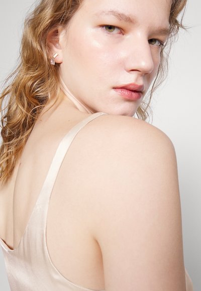 Σιφόν top σε μπεζ απόχρωση, με λεπτές τιράντες, απαλό υλικό και διακριτική γυαλάδα. Τα κυματιστά μαλλιά του μοντέλου και τα χρυσά σκουλαρίκια προσθέτουν λεπτομέρεια.