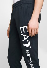 Melnas sporta bikses ar mīkstu tekstūru, ar baltu "EA" logotipu un "EMPORIO" tekstu sānos, elastīgu jostu un sānu kabatām.