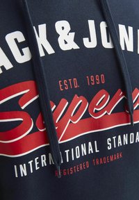 Navy Hoodie mit auffälligem weißen und roten Text. Das Design enthält "Super" in großer Schreibschrift, sowie "ESTD. 1990" und "INTERNATIONAL STANDARD."