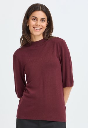 Jeune femme souriante, portant un pull bordeaux à manches courtes et un pantalon noir, debout devant un fond clair uni.