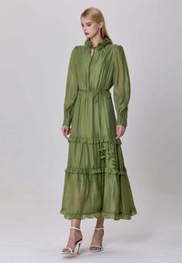 Femme portant une longue robe verte olive à volants avec des détails de col floral et des sandales à talons blancs, se tenant devant un fond uni.