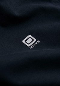 Tessuto nero con logo quadrato bianco e testo "ambre" stampato sulla superficie, mostrando la texture e le pieghe del tessuto.