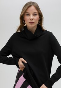 Svart stickad högkragströja med långa ärmar och en lös krage. Slät textur, figursydd design, bärs med ett plagg i rosa och svarta ränder.