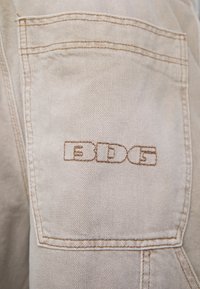 Beige denimjacka med ficka och bruna sömmar, prydd med en rektangulär broderad logotyp "EDG" i ett fet, modernt typsnitt.