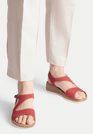 Pies con pantalones beige y sandalias de cuña abiertas en rojo con tiras en el tobillo y suelas blancas sobre un fondo claro.