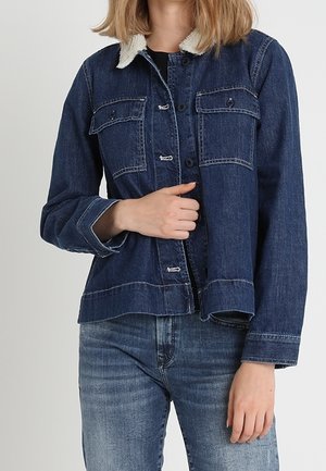 Personne portant une veste en jean bleu foncé avec un col en peau lainée blanche et un jean bleu délavé, tenant la veste près de la taille.