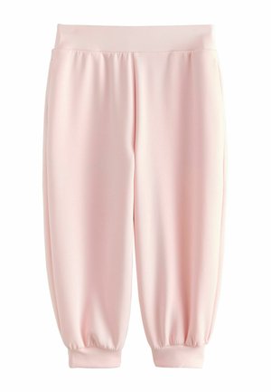 Persoană purtând pantaloni scurți roz deschis cu tivuri decupate și adidași albi, stând în fața unui perete simplu și deschis la culoare.