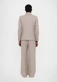 Šviesiai pilkas patikimo rašto kostiumas, turintis pritaikytą švarką ir plačias kelnes. Išskirtinė rožinė ir balta tinklelio potėpė, tekstūruota medžiaga ir tvirta siluetas.