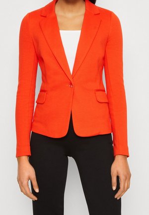 Kobieta ubrana w dopasowaną, jasno pomarańczową marynarkę na białą bluzkę i czarne spodnie, widziana od szyi do bioder z ramionami swobodnie opuszczonymi wzdłuż ciała.