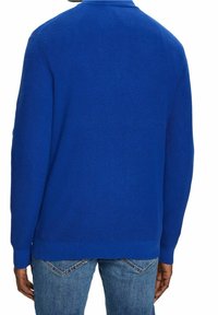 Personne vue de dos portant un pull en maille texturée bleu vif et un jean bleu moyen avec poches arrière visibles.