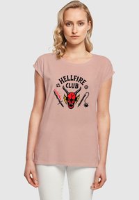 Camiseta de corta manga en rosa claro con un gráfico del "Hellfire Club" que presenta la cara de un demonio rojo, dos armas y elementos decorativos en negro.
