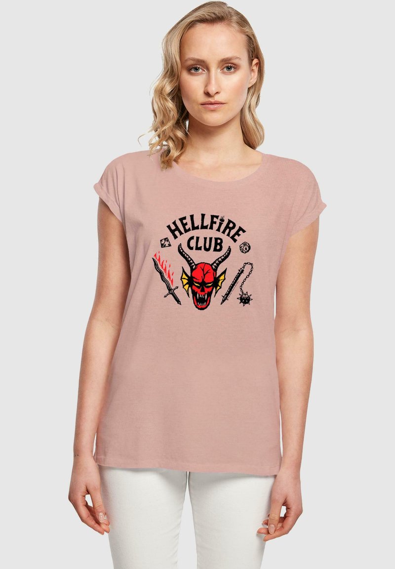 Camiseta de corta manga en rosa claro con un gráfico del "Hellfire Club" que presenta la cara de un demonio rojo, dos armas y elementos decorativos en negro.