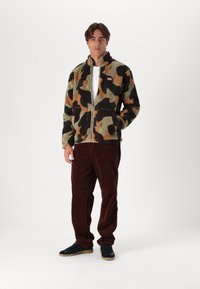 Giacca in pile mimetica nei colori verde, marrone e nero, con colletto alto, zip frontale e due tasche laterali, abbinata a pantaloni in corduroy marroni e scarpe nere.