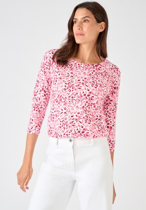 Vrouw gekleed in een roze- en roodgestreepte top met lange mouwen en een witte broek met hoge taille, staand tegen een effen achtergrond.