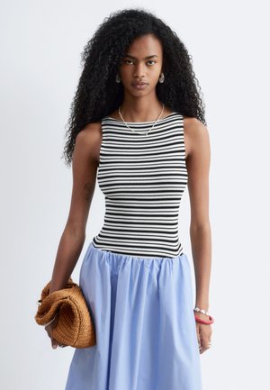 Femme aux longs cheveux bouclés porte un top sans manches rayé noir et blanc, une jupe bleu clair froncée, tenant une pochette tressée couleur camel.