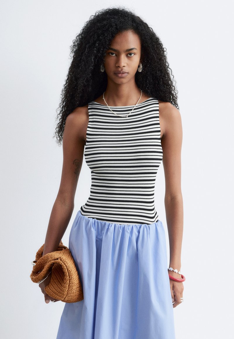 Femme aux longs cheveux bouclés porte un top sans manches rayé noir et blanc, une jupe bleu clair froncée, tenant une pochette tressée couleur camel.