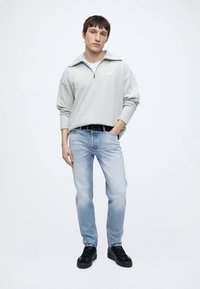 Jeune homme debout, une main dans la poche, portant un pull gris clair à fermeture éclair quart, un jean bleu clair et des baskets noires sur fond blanc.