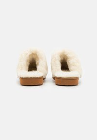 Des chaussons moelleux de couleur crème avec une texture douce, présentant un design à dos ouvert et une semelle marron clair.
