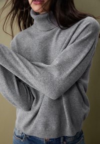 Ženska oblečena v svetlo sivo turtleneck pulover z dolgimi rokavi, prekrižanimi čez prsi, v kombinaciji z modrimi kavbojkami, na zatemnjenem bež ozadju.