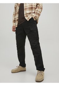Black cargo pants with multiple side pockets, crafted from durable cotton. Worn with a beige flannel shirt and light-colored shoes.