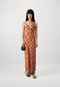 Maxi dress in brown and orange plaid pattern, with a keyhole detail and adjustable straps. Sheer hem. Model holds a small black handbag.