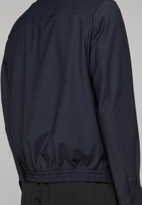 Giacca blu navy con elastico in vita indossata da una persona, vista posteriore e parte di pantaloni neri.