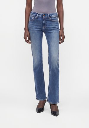 Femme portant un jean bleu délavé à jambes droites, un haut gris rentré et des talons noirs pointus, debout sur un fond blanc.