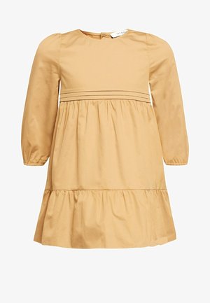 Kind draagt een lange mouw beige jurk met een geplooide taille en zoom, staat blootsvoets tegen een neutrale achtergrond.