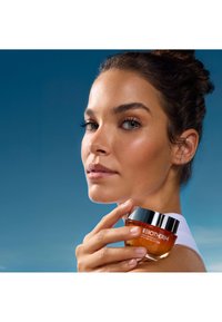 Pomarańczowy żel krem Biotherm z czarną pokrywką, trzymany przez osobę, na niebieskim tle. Skóra wydaje się gładka i zdrowa.