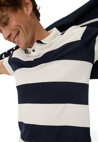Navy and white striped polo shirt with a collar and two buttons, featuring a soft texture. Dark sweater draped over the shoulder.