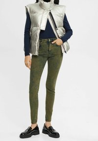 Chaleco acolchado plateado metálico con cuello alto, combinado con jeans ajustados verde oscuro y loafers de plataforma negros. Textura suave y diseño elegante.