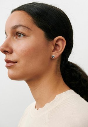 Ung kvinde med brunt hår trukket tilbage, iført en cremefarvet top og små sølv hjerteformede ørestikker, kigger til venstre mod en ensfarvet baggrund.