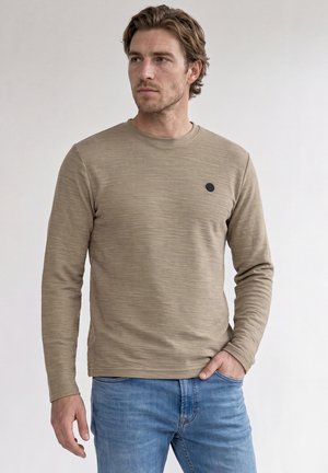Jonge man die een beige longsleeve overhemd en blauwe spijkerbroek draagt, staand met één hand in zijn zak, zijwaarts kijkend tegen een effen achtergrond.
