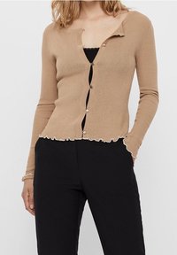 Cardigan beige a coste con maniche lunghe, orlo ondulato e cinque bottoni. Indossato sopra una maglietta nera, abbinato a pantaloni neri.