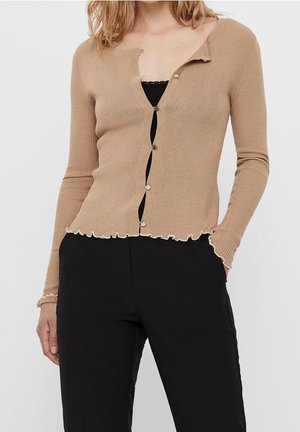 Cárdigan de color beige con canalé, de mangas largas, con dobladillo de picos y cierre de cinco botones. Lleva puesto sobre una blusa negra, combinada con pantalones negros.