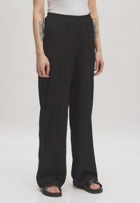 Schwarze, weite Hosen aus ribbeliger Textur mit elastischem Bund und seitlichen Taschen, kombiniert mit flachen schwarzen Sandalen vor einem neutralen Hintergrund.
