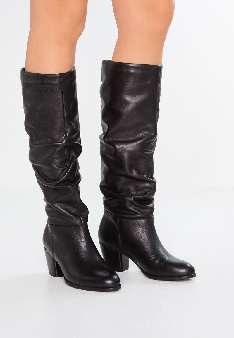 Bottes en cuir noires jusqu'au genou avec un design décontracté, un talon bloc et une texture lisse. Présente une pointe arrondie et des détails minimaux.