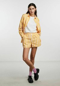 Gul og hvid stribet skjorte med knapper og matchende shorts. Hvid t-shirt under, sorte sko og lyserøde sokker. Glat tekstur.