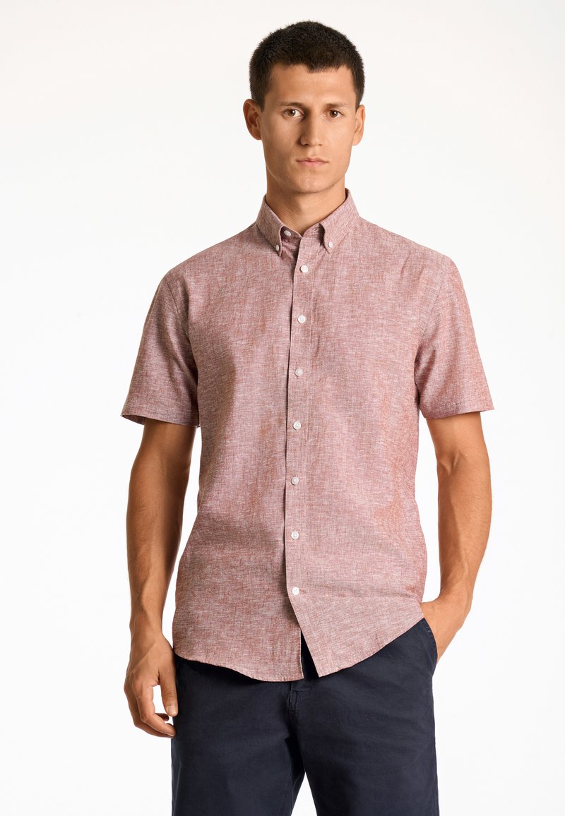 Man in een kortearm, knoopshirt met een roze textuur en donkerblauwe broek, staand met één hand in de zak tegen een witte achtergrond.