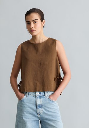 Junge Frau mit dunklen Haaren, trägt ein braunes ärmelloses Oberteil mit seitlichen Bändern und hellblaue, hoch taillierte Jeans, Hände in den Taschen.