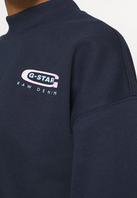 Mörkblå crew-neck sweatshirt tillverkad av mjukt tyg, med en flerfärgad G-Star Raw Denim-logotyp på vänster bröst.