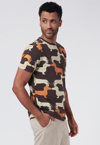 Kortärmad t-shirt med en brun bakgrund och ett repetitivt mönster av dachshundsilhuetter i beige och orange. Mjuk bomullstyg.